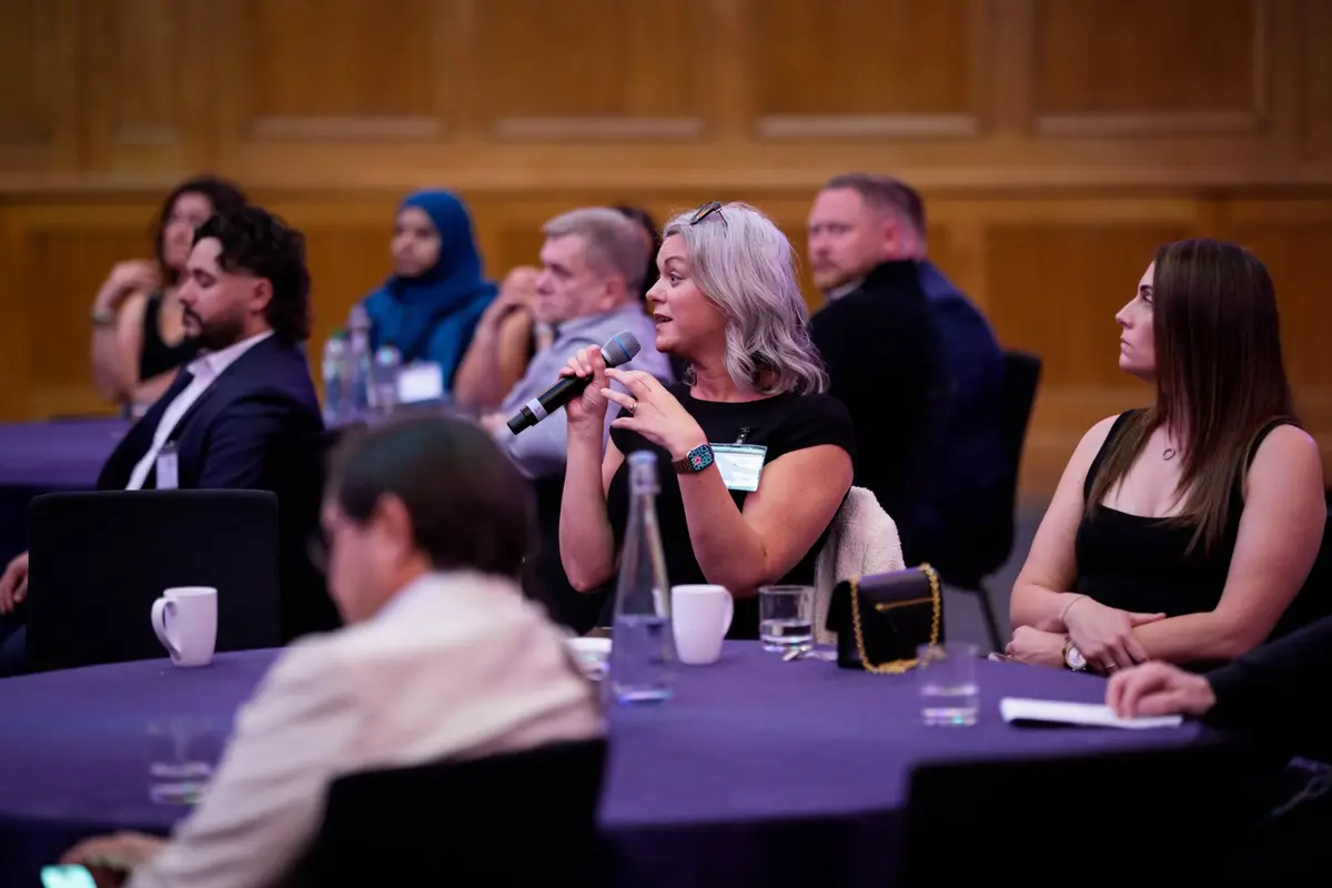 A delegate speaking into a handheld microphone during an open discussion at a conference, with other delegates listening from round tables.
