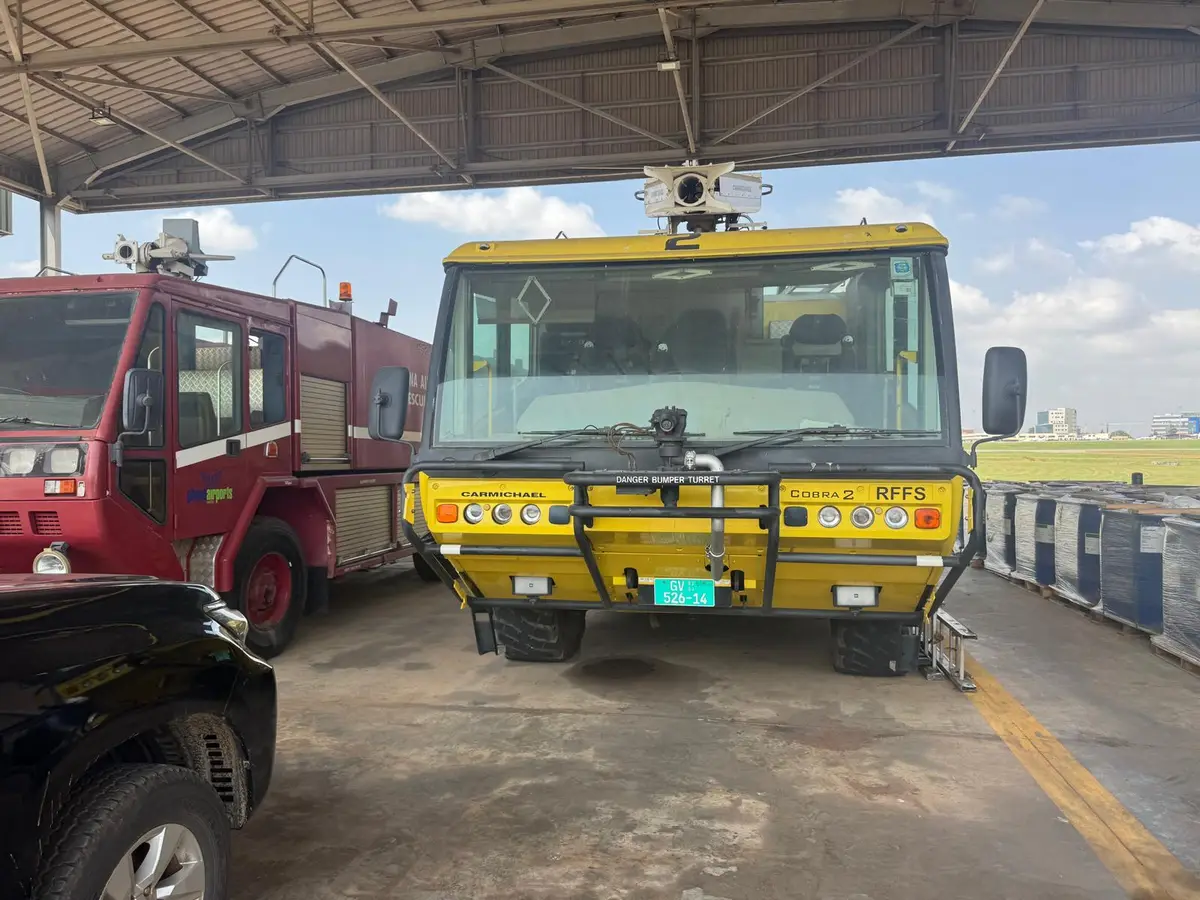Two airport rescue and firefighting appliances parked under an open vehicle shelter, with the airfield visible beyond.